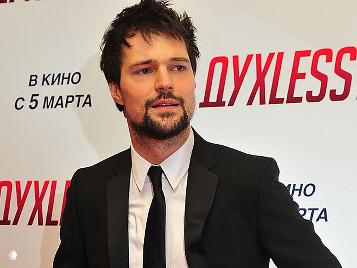 Мужской портрет в костюме и галстуке на красной дорожке перед баннером с логотипом фильма «ДУХLESS», официальный кадр с презентации фильма.