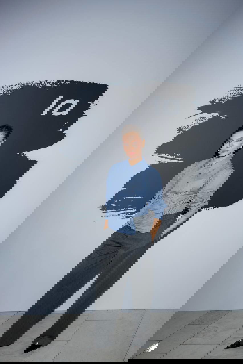 Строгий повседневный образ: светло‑голубая рубашка и тёмные брюки на фоне графического баннера в интерьере галереи после показа Lamoda.