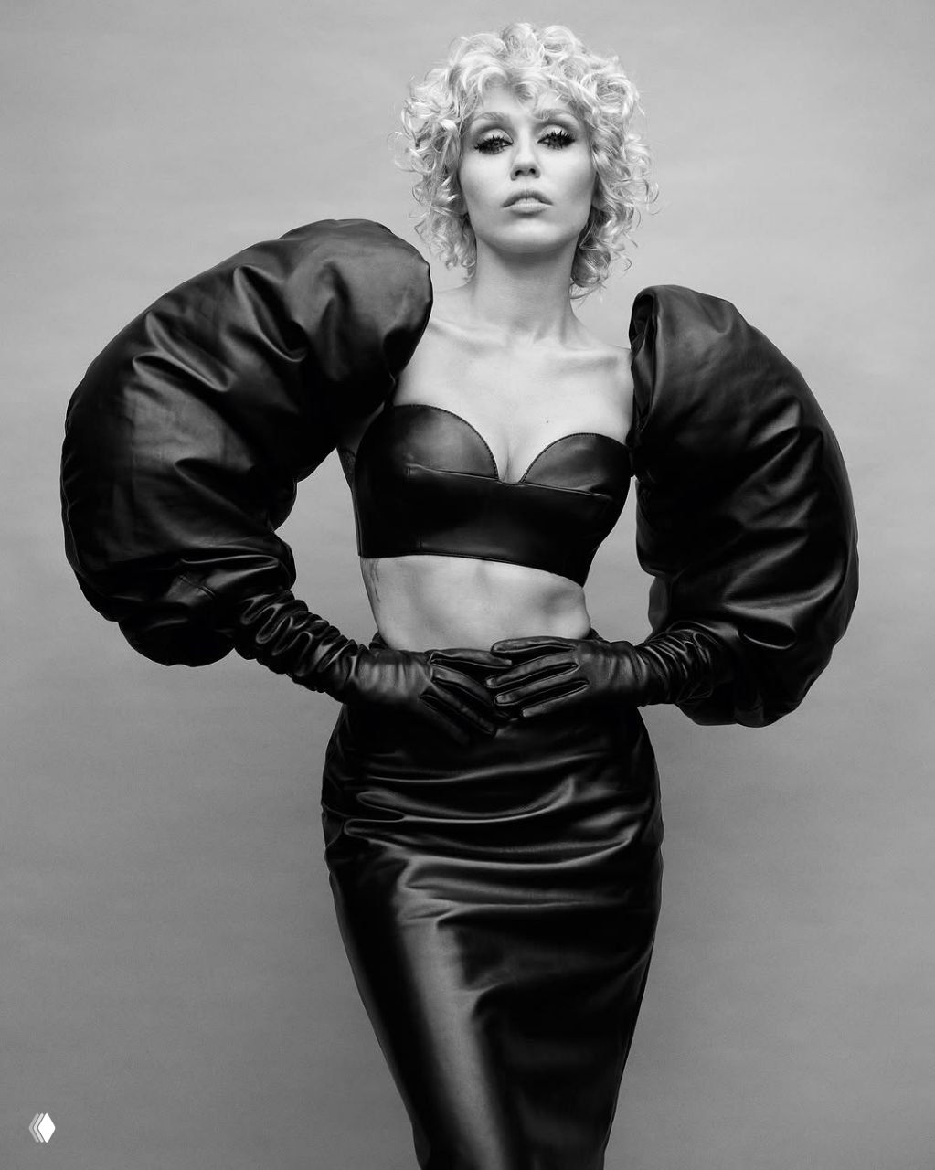 Майли Сайрус в чёрно‑белой студийной съёмке: кожаный бра, массивные пышные рукава и уверенная поза в стилизованном гардеробе.
