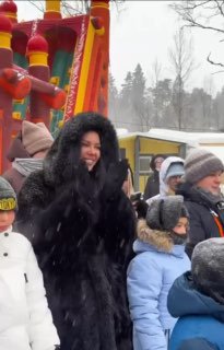 Оксана Самойлова в тёмной шубе улыбается и аплодирует на масленичных гуляньях на ферме, вокруг дети в зимней одежде и снег.