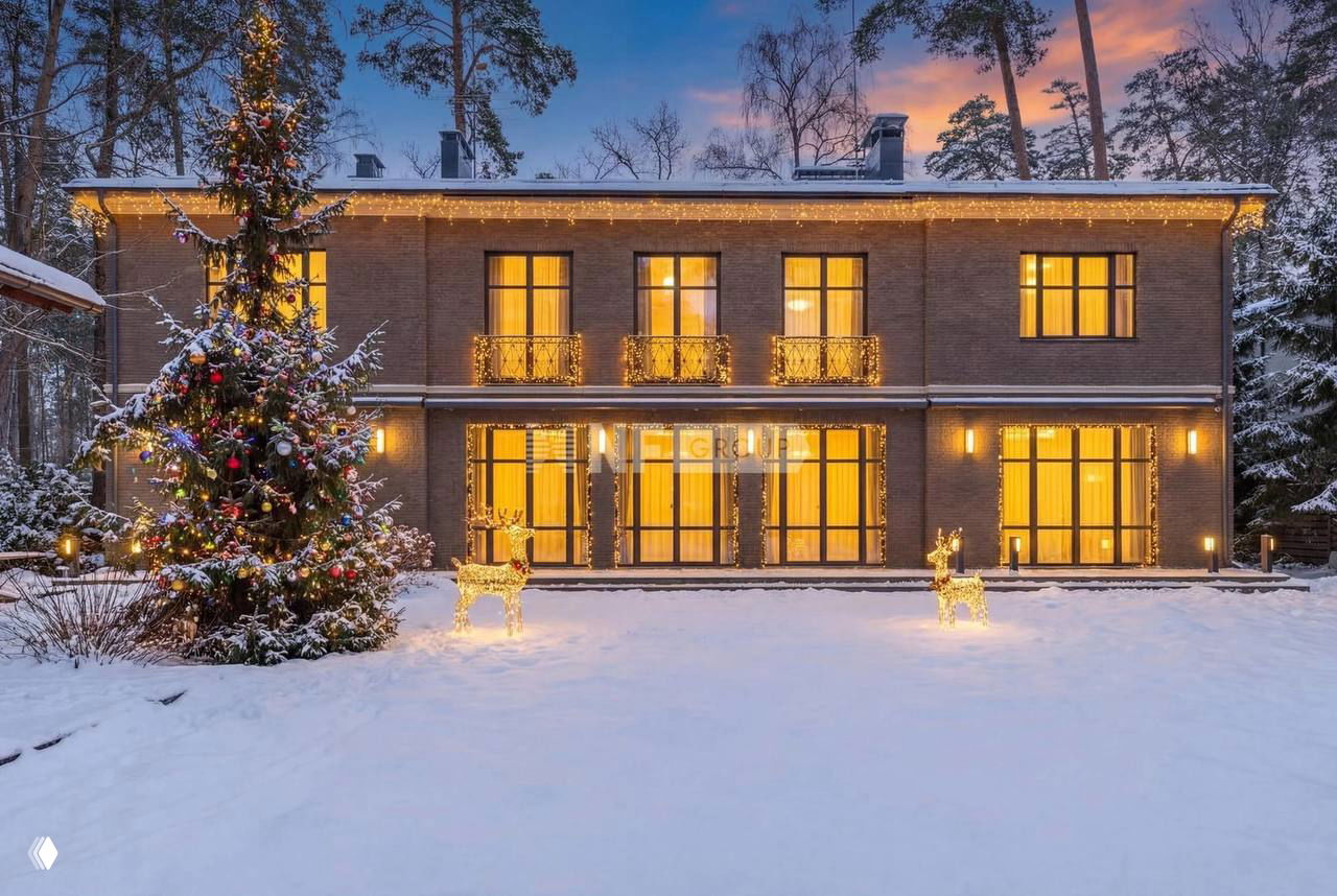 Фасад двухэтажного кирпичного особняка на участке, зимний пейзаж со снегом, рождественская ёлка и декоративная подсветка вечером.
