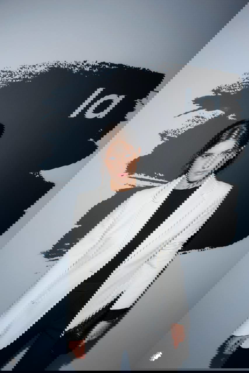 Женщина в белом пиджаке и чёрном образе смотрит в камеру на фоне большого логотипа и графического пятна — минималистичный лук мероприятия.