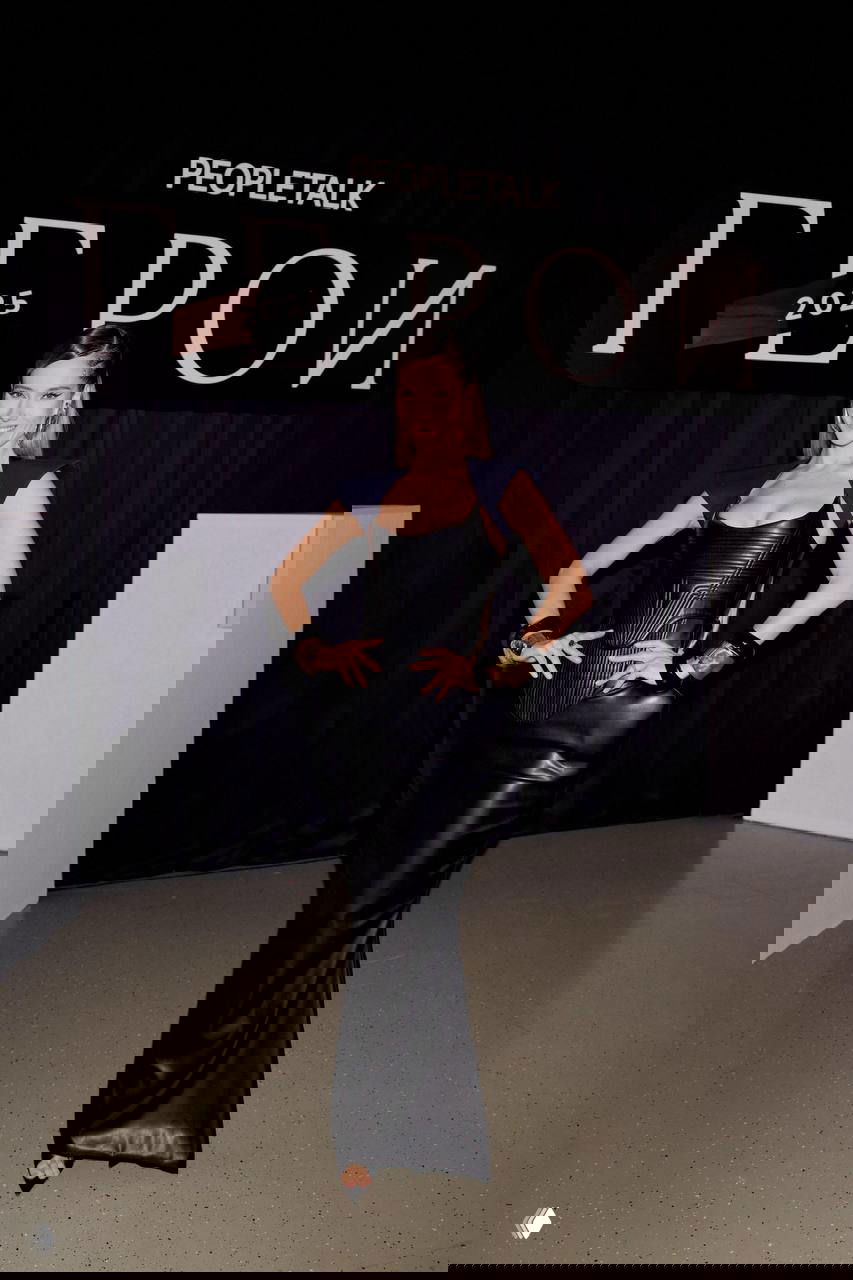 Гостья в обтягивающем чёрном платье с корсетным верхом и крупными браслетами позирует у брендированного фона события — модный образ вечера.