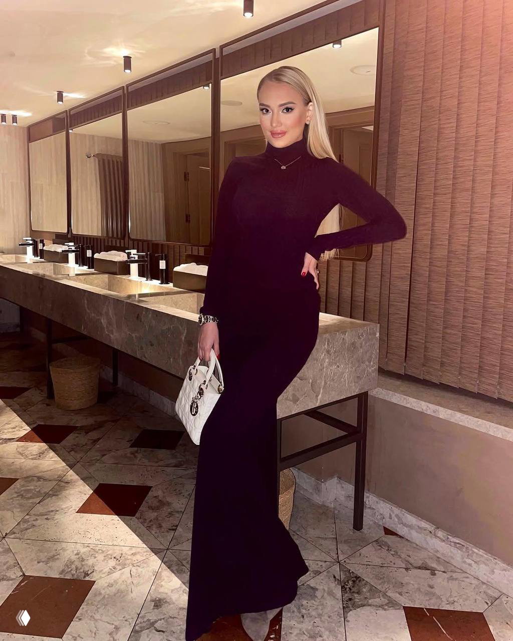 Ксения Гнедина в обтягивающем длинном чёрном платье в интерьере общественного пространства с зеркалами и столами, вечерний элегантный образ в полный рост.