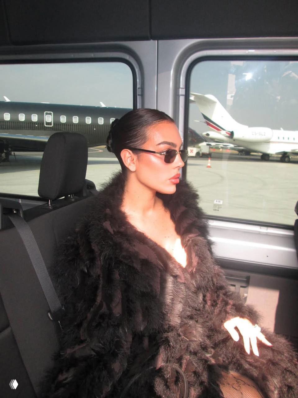 Крупный план Джорджины Родригез в шубе и солнцезащитных очках в салоне автомобиля на фоне частных самолётов у терминала аэропорта.