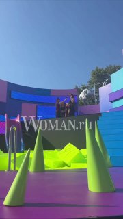 Яркая сцена подиума с цветными фигурами и конусами; на дорожке видна женщина с детьми, заметен водяной знак Woman.ru внизу кадра.