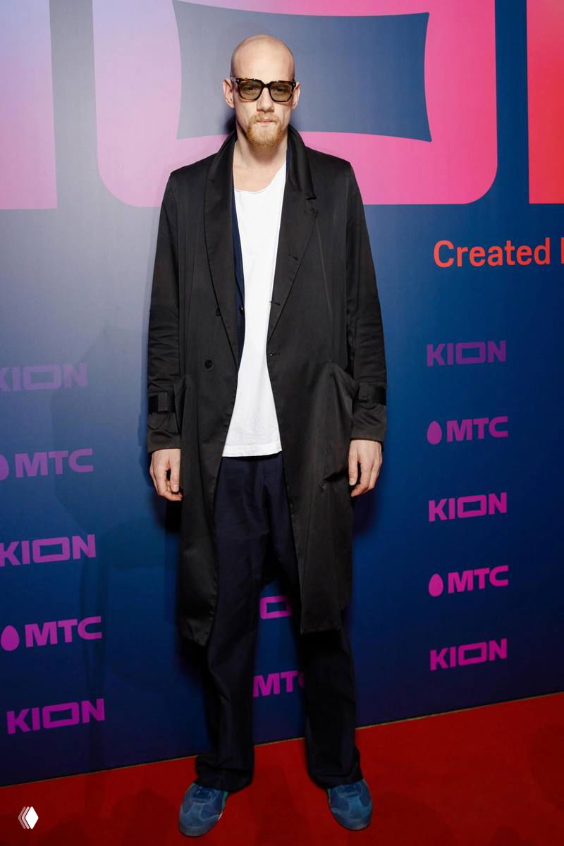 Мужчина в длинном пальто и очках позирует на красной дорожке у бренд-стены мероприятия в затемнённом помещении, журналистские кадры.