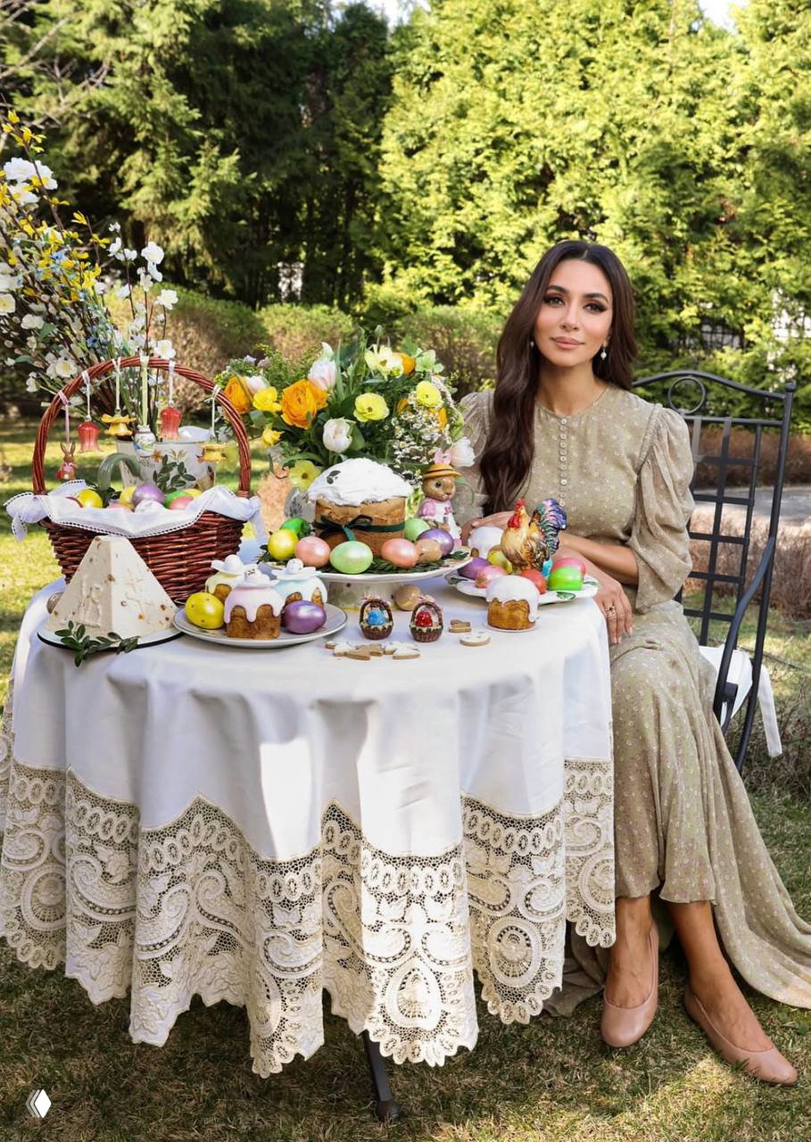 Женщина в платье сидит за садовым столиком с богато украшенным пасхальным столом: разноцветные яйца, куличи, цветы и праздничная посуда на природе.