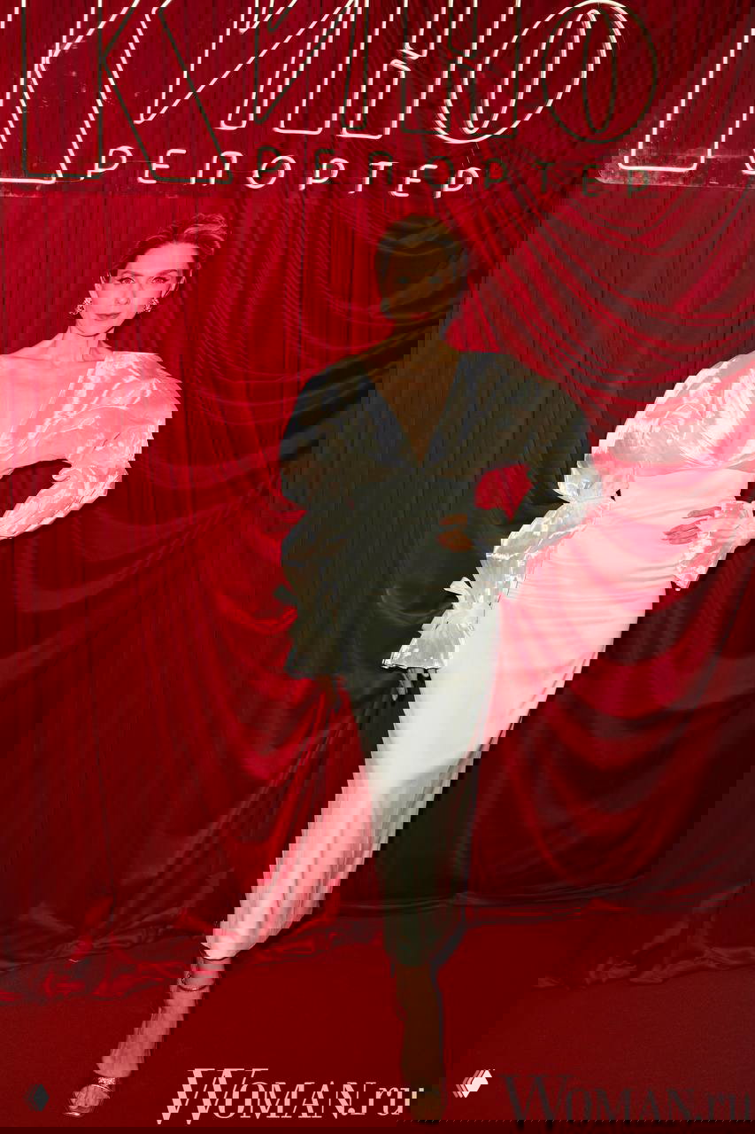 Женщина в атласном белом платье с объёмными рукавами позирует на фоне красной драпировки и неоновой вывески «КиноРепортер».