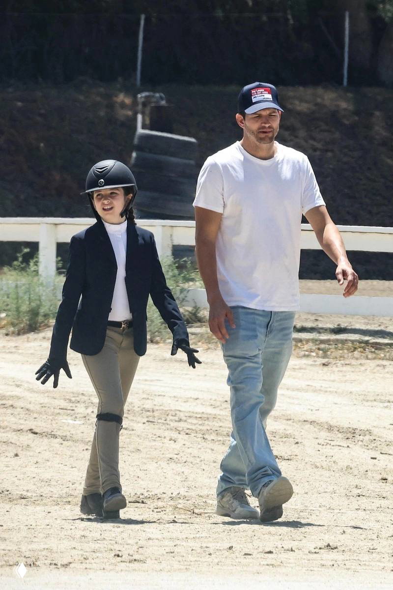 Эштон Кутчер идёт рядом с дочерью, одетой для верховой езды; снимок на загородной площадке показывает прогулку и экипировку.