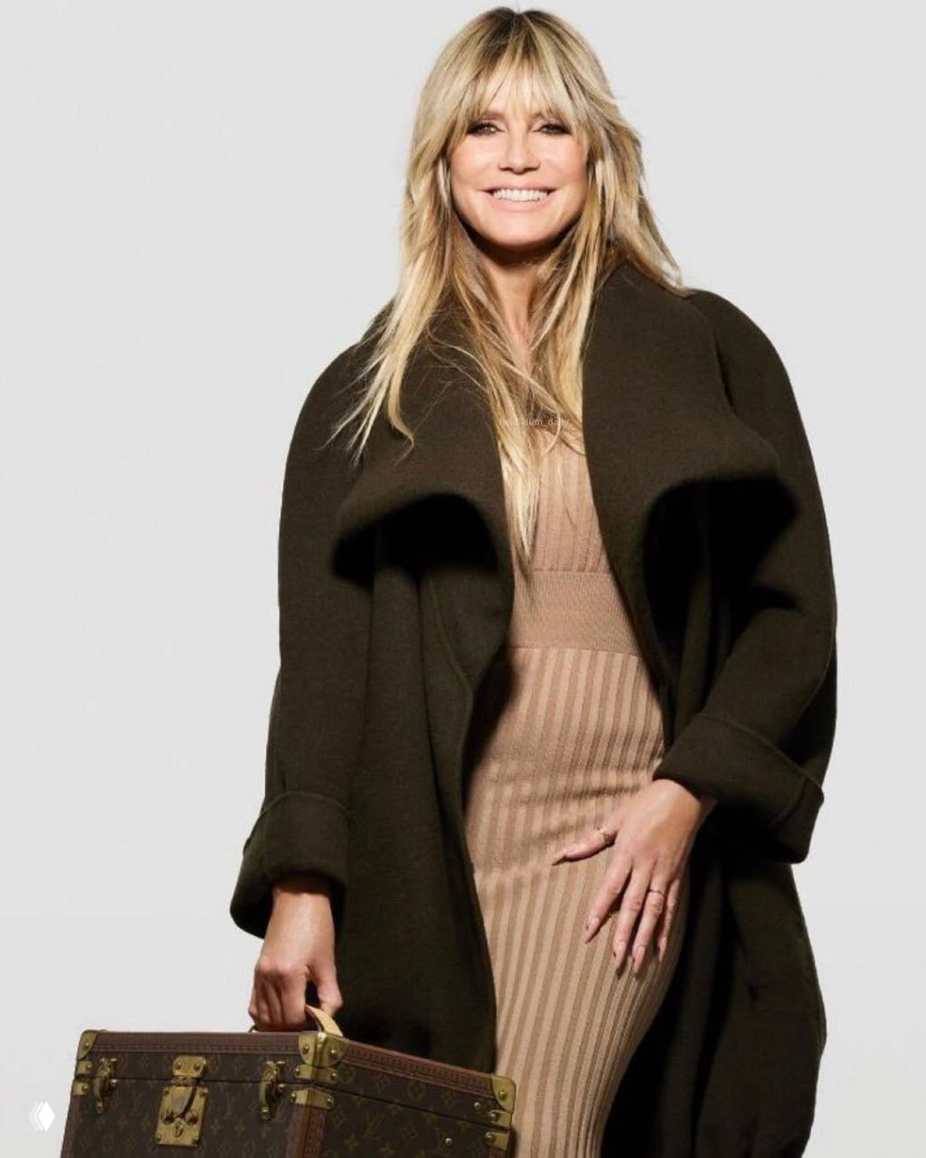 Фотография в полный рост: женщина в тёплом пальто и облегающем платье держит дорожный чемодан в кадре модной съемки на сером фоне.