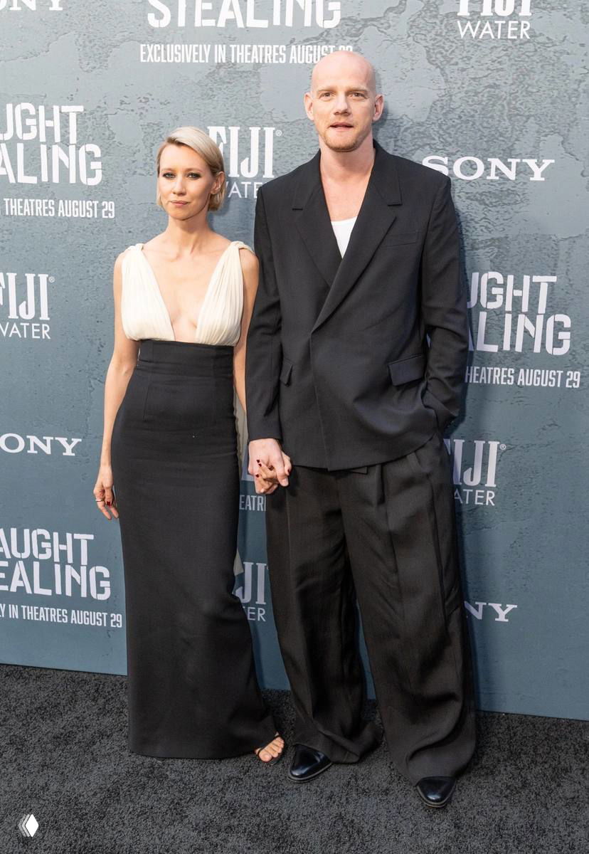 Пара держится за руки на премьере: мужчина в чёрном пиджаке и женщина в вечернем платье на фоне пресс-волла кинофильма.