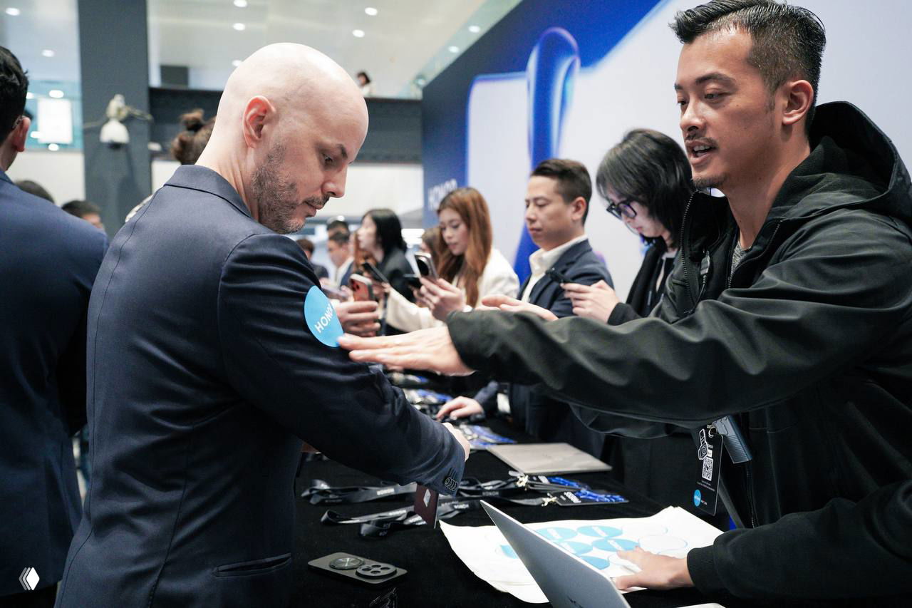 Посетители на выставочном стенде Mobile World Congress: сотрудник помогает гостю у стола с устройствами, видны демонстрационные аксессуары.