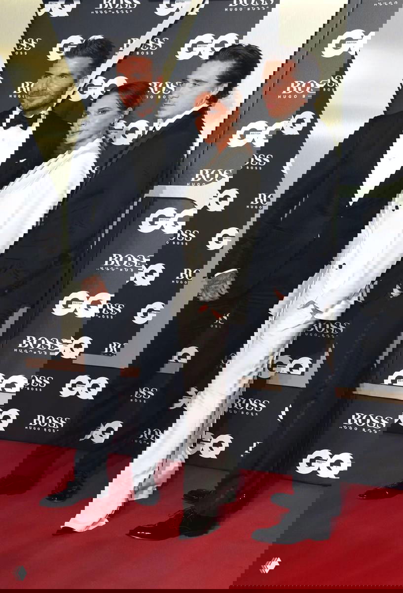 Трое людей на красной дорожке перед логотипами события: двое мужчин в смокингах и женщина в светлом костюме, позируют для фотографов.