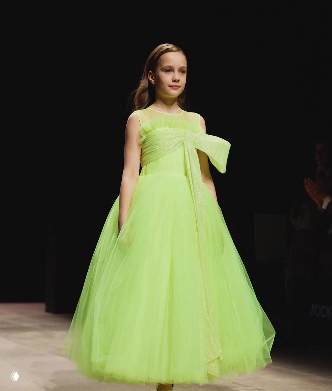 Девочка идёт по подиуму в салатовом пышном платье на показе Московской недели моды; чёрный фон подчёркивает образ и свет на модели.