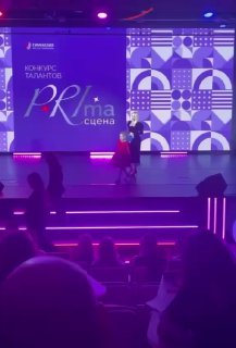 Широкий кадр сцены с цветной подсветкой и девочкой в центре, выступление на конкурсе талантов Prima сцена, зрители в затемнённом зале