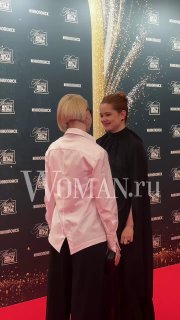Мила Ершова в черном платье общается на красной дорожке премии АПКиТ; рядом человек в светлой рубашке, видны логотипы и фон.