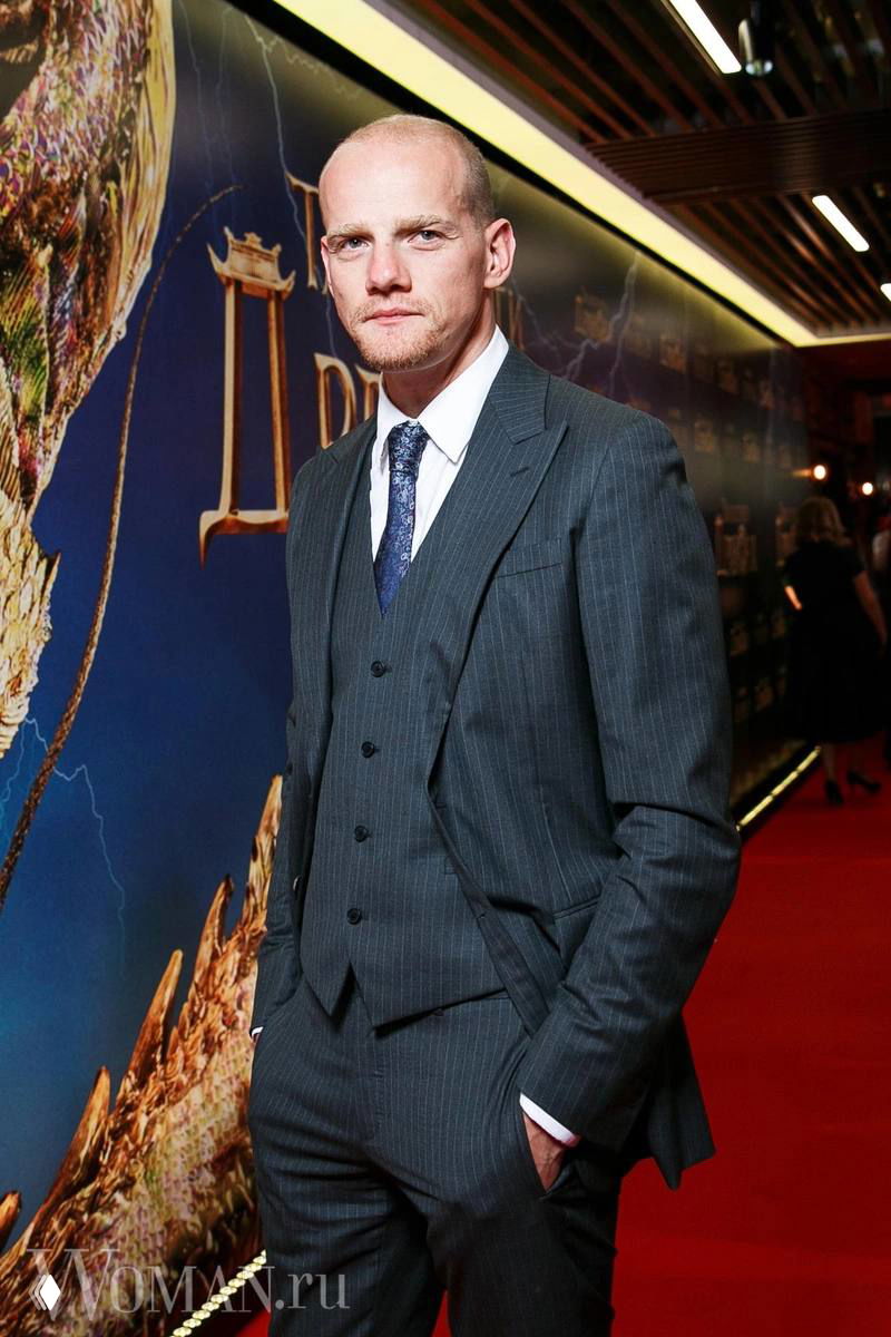 Портретный снимок мужчины в строгом костюме на фоне постера мероприятия, кадр с премьеры с официальной фотосессии.