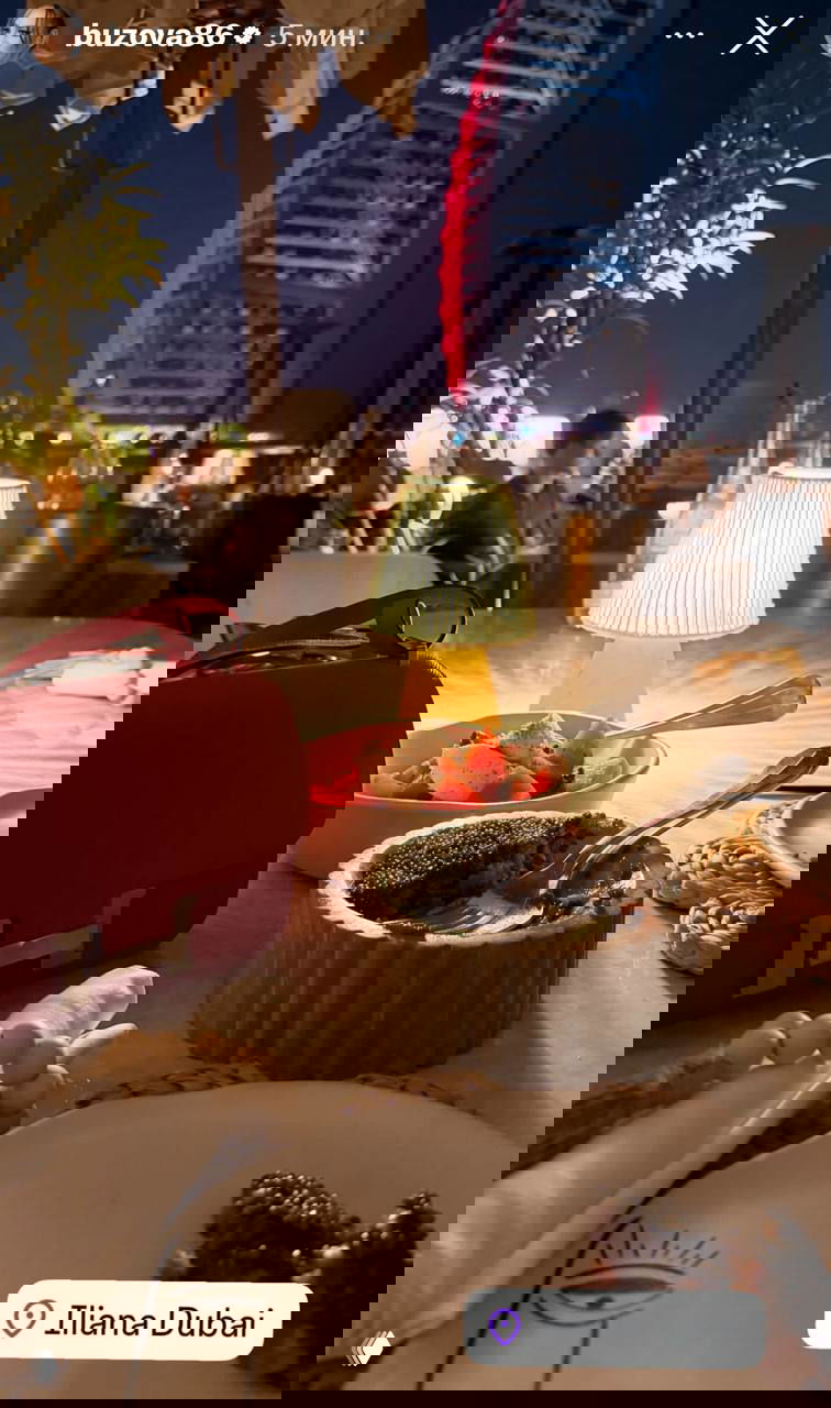 Ночной стол в ресторане с десертами и салатом, яркая красная сумка на переднем плане; вдалеке виден подсвеченный силуэт отеля Burj Al Arab.