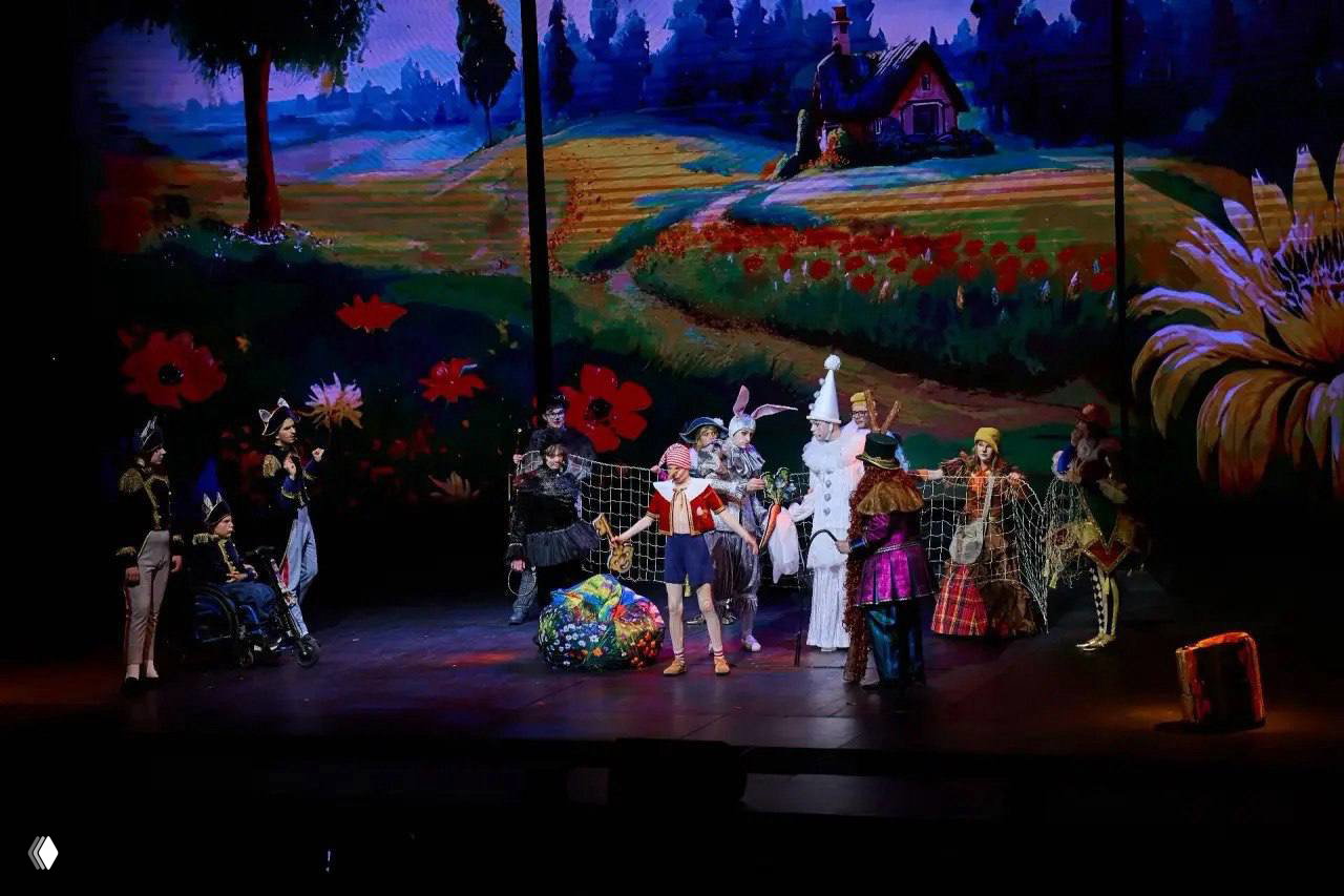 Широкий план сцены с коллективом актёров инклюзивной труппы, яркие декорации и костюмы в массовой сцене спектакля.