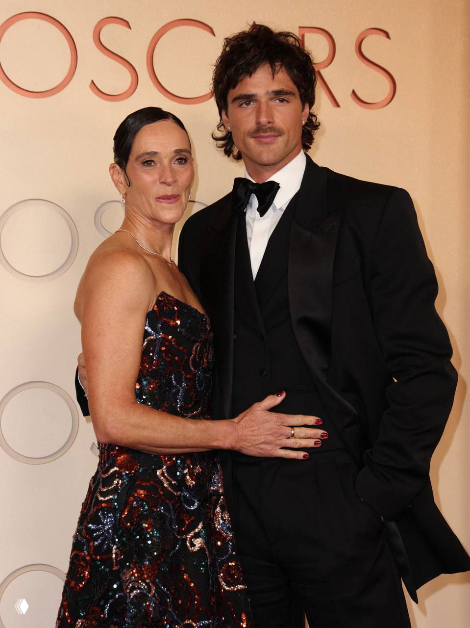 Крупный план пары на церемонии: мужчина в смокинге и женщина в пайетчатом платье позируют рядом с логотипом «Oscars» на фоне пресс-волла.