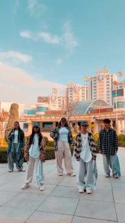 Группа молодых людей на открытой городской площадке у современного комплекса, выстроены в ряд и позируют/двигаются в танце на фоне архитектуры