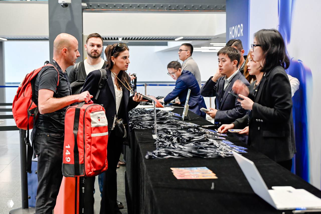 Посетители у стола выдачи на стенде Honor: люди с багажом, представители компании и стол, заваленный лентами и бейджами.