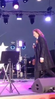 Мужчина идёт по сцене с микрофоном; на заднем плане видны колонки, световые приборы и сценическое оборудование во время выступления на площадке.