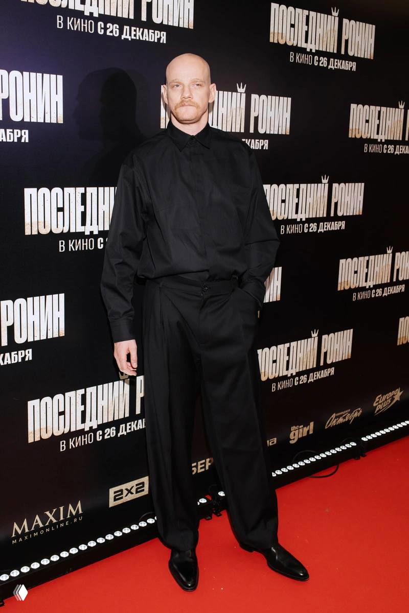 Мужчина в чёрной рубашке и брюках позирует на красной дорожке у афиши фильма; светский снимок с премьеры в кинотеатре.