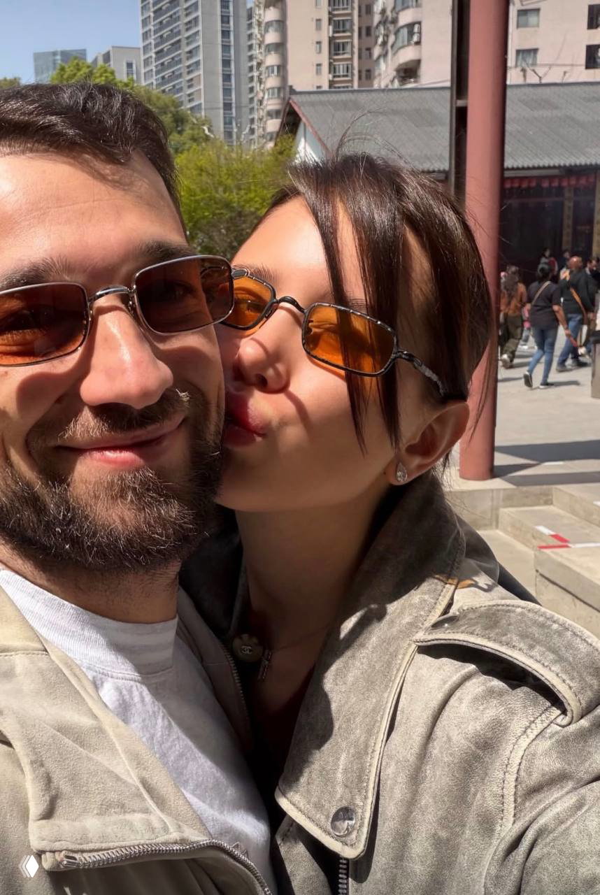 Аврора Киба и мужчина (предположительно Рустам Гаджиев) обнимаются и позируют для селфи в солнечных очках на фоне городской застройки и прохожих.