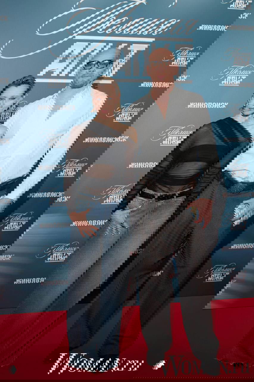 Дуэт на красной дорожке: мужчина в белой рубашке и очках с женщиной в чёрном наряде на фоне баннера церемонии награждения.