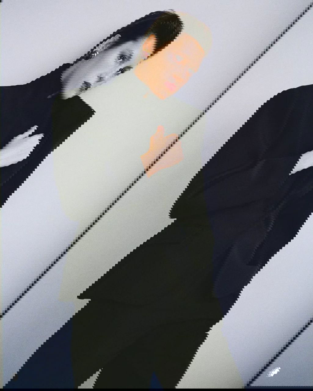 Женщина в тёмном жакете и свободных брюках на светлом фоне; сдержанная постановочная фотография с акцентом на силуэт и детали.