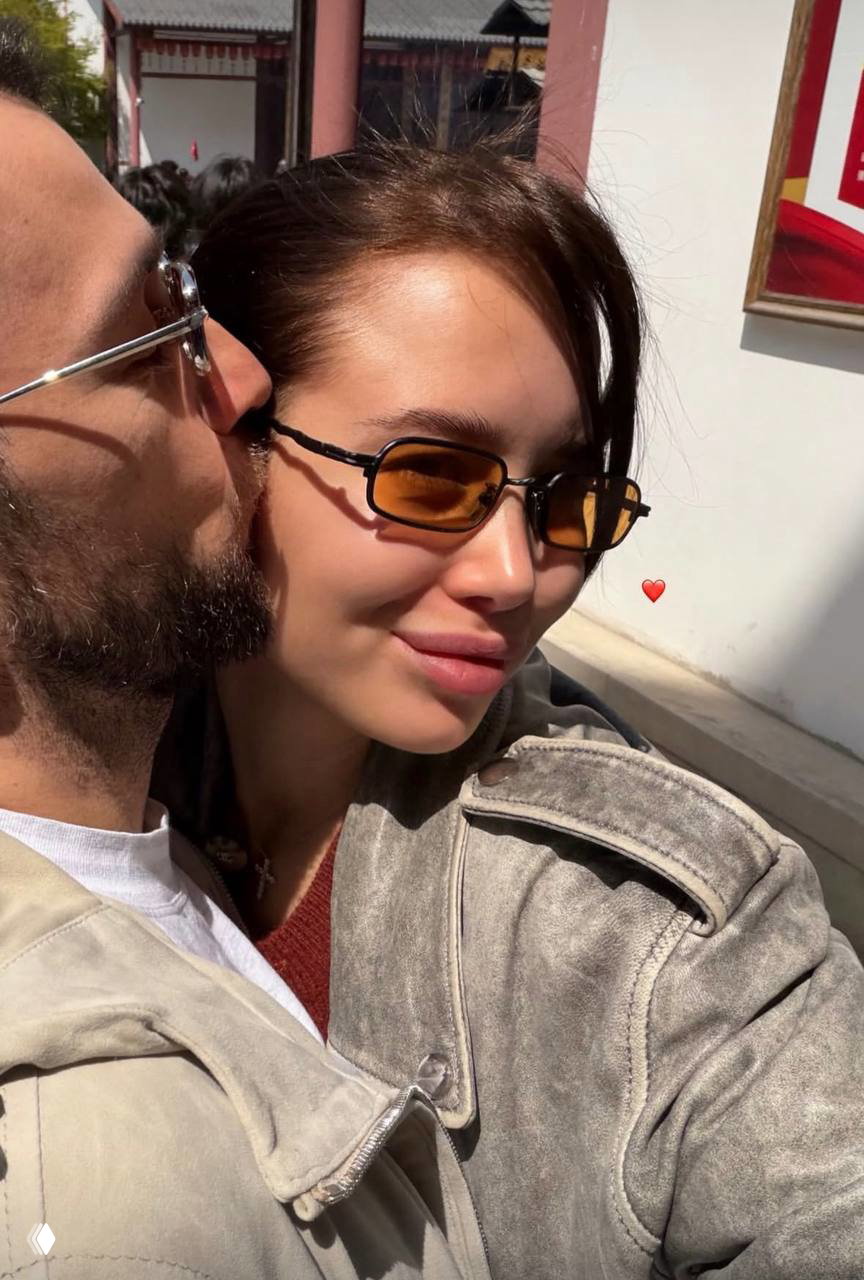 Крупный план пары: женщина принимает поцелуй в щёку от мужчины, они в солнечных очках и куртках, на заднем плане видна туристическая локация и люди.