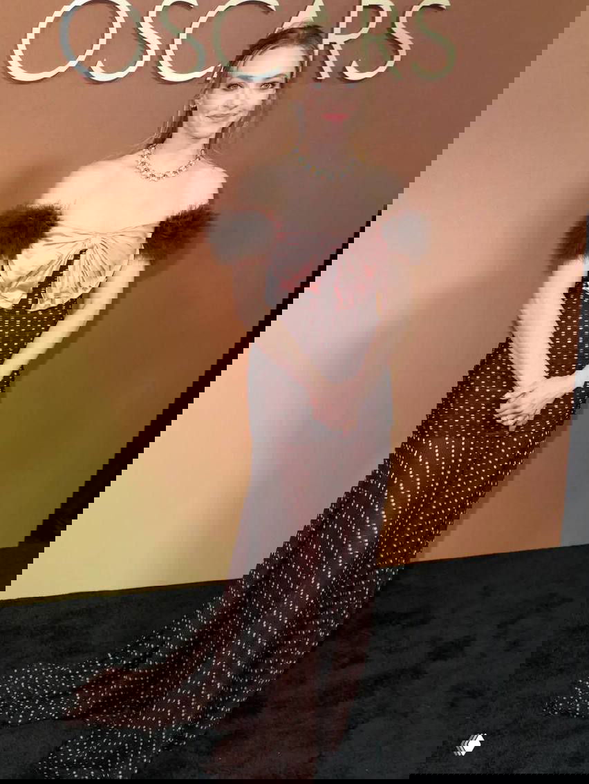 Женщина в длинном коричневом платье с бантовой декоративной деталью и открытыми плечами, кадр на фоне композитного декора OSCARS.