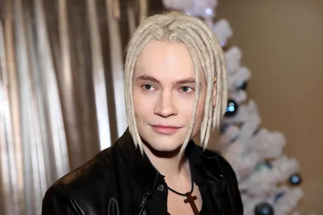 Портрет певца Шамана в светлых дредах и кожаной куртке, он улыбается на фоне украшенной новогодней ёлки в помещении.