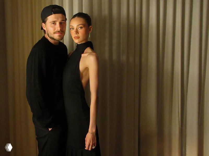 Никола Пельтц в длинном чёрном платье в полубоковом ракурсе на фоне штор; кадр с цифровыми артефактами, праздничная обстановка