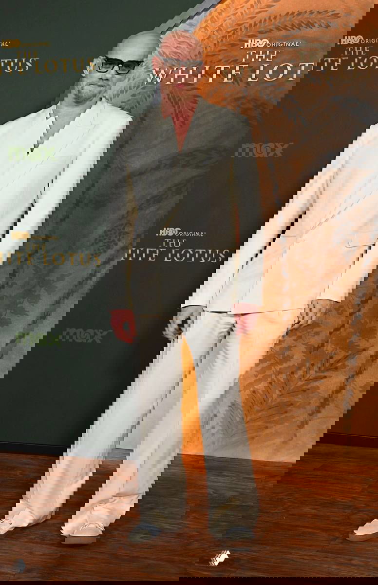 Мужчина в светлом костюме позирует на пресс-волле сериала; фото с премьеры телепроекта в интерьерной обстановке.