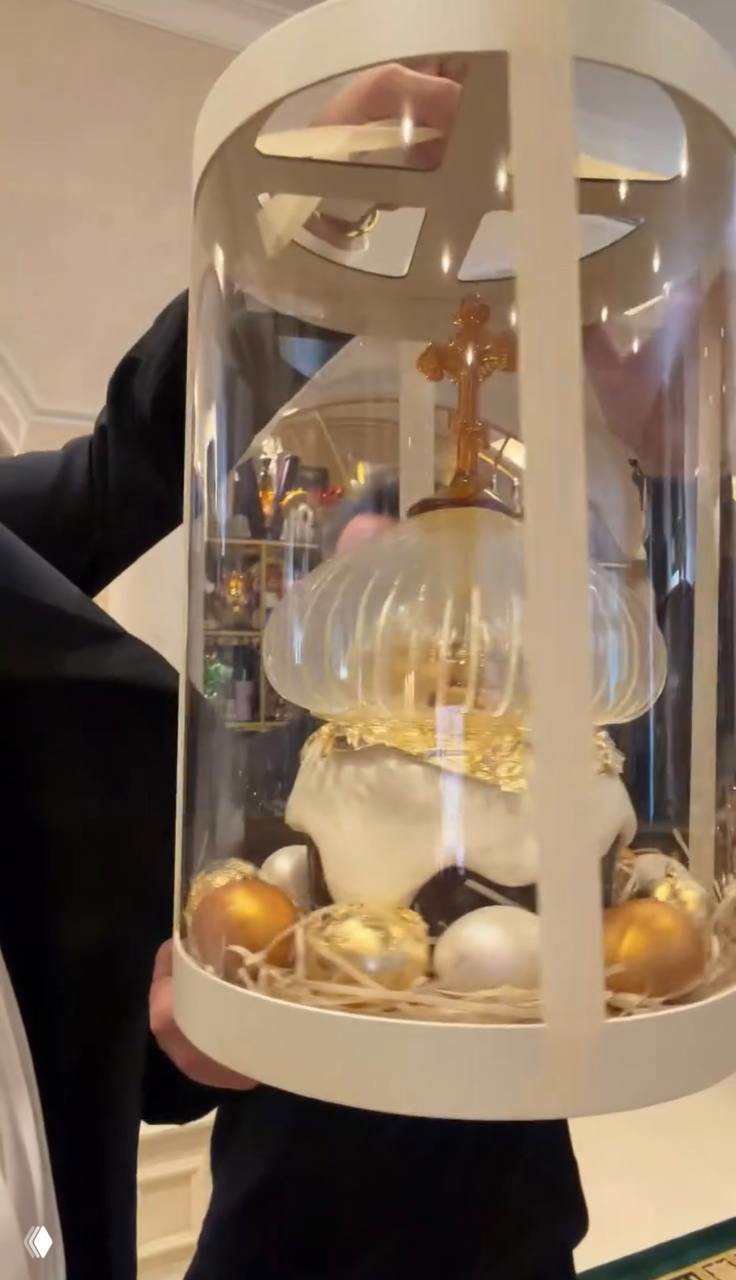 Крупный план подарочного пасхального набора в прозрачной декоративной колбе: украшенный кулич в оформлении с золотыми шарами и лентой.