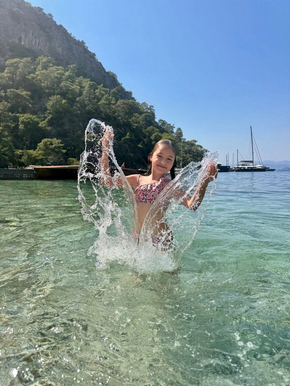 Девочка в купальнике плескается в прозрачной прибрежной воде, на заднем плане яхты и лесистый берег, летняя морская сцена