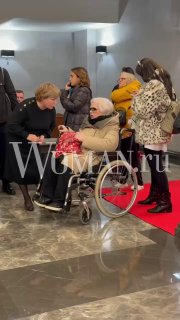 Светлана Крючкова в инвалидной коляске среди гостей в фойе, рядом люди общаются, фотографы и красная дорожка видны на заднем плане.