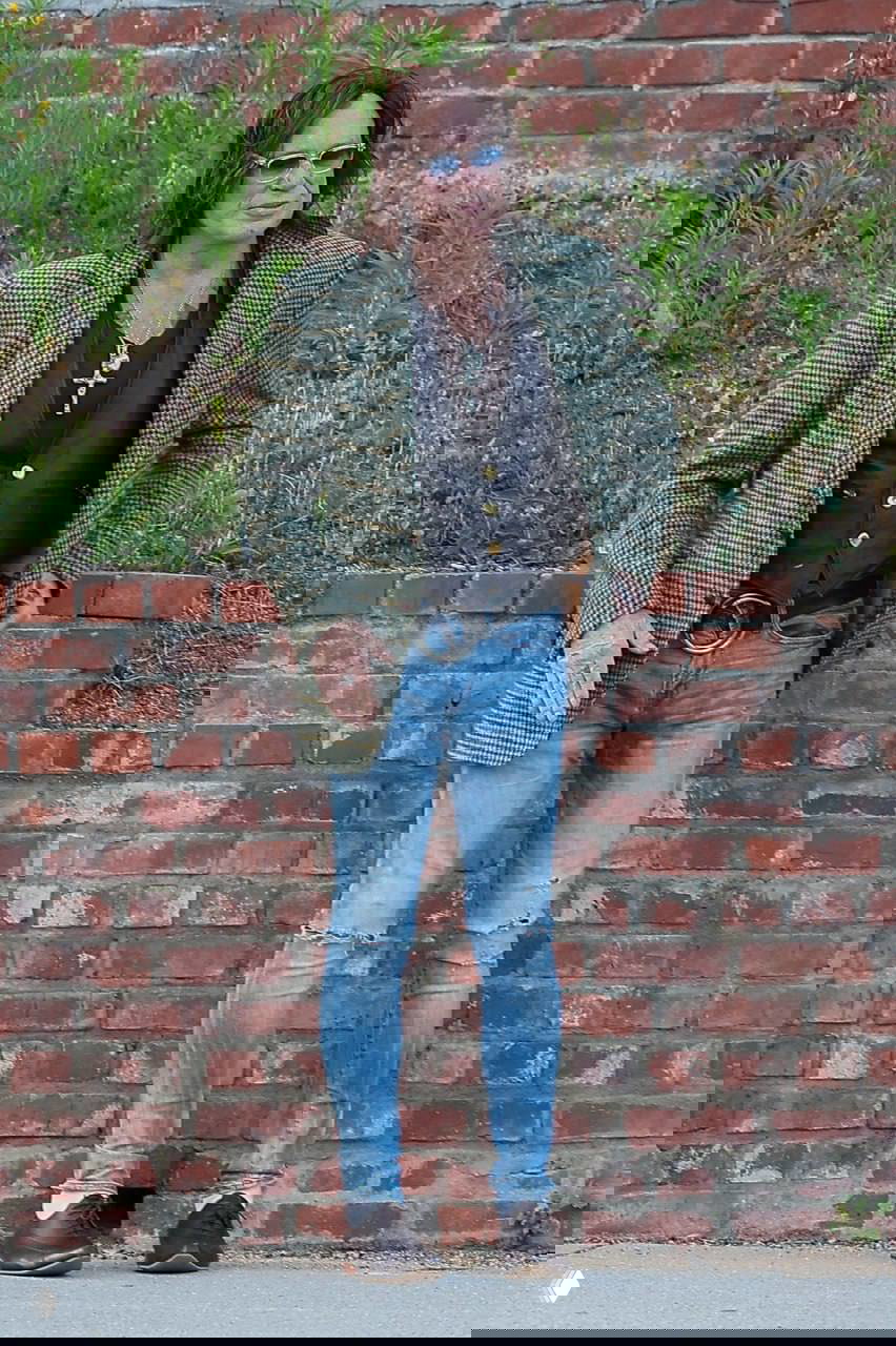 Кадр крупным планом: Микки Рурк позирует в скинни и пиджаке на фоне зелени у кирпичного забора в Нью‑Йорке.