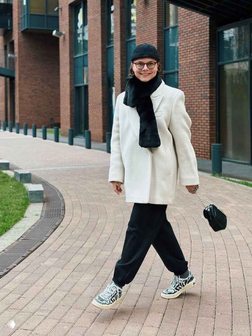 Татьяна Брухунова в светлом пальто и тёмном шарфе идёт по тротуару в городской застройке, в удобных кедах и с мини‑сумкой.