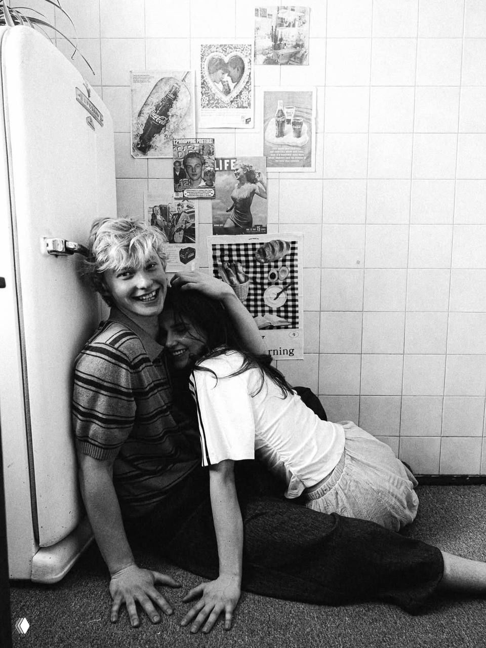 Чёрно‑белый кадр в кухонном пространстве: Полина Денисова и Антон Рогачёв сидят у холодильника, смеются, на стене видны вырезки и постеры.