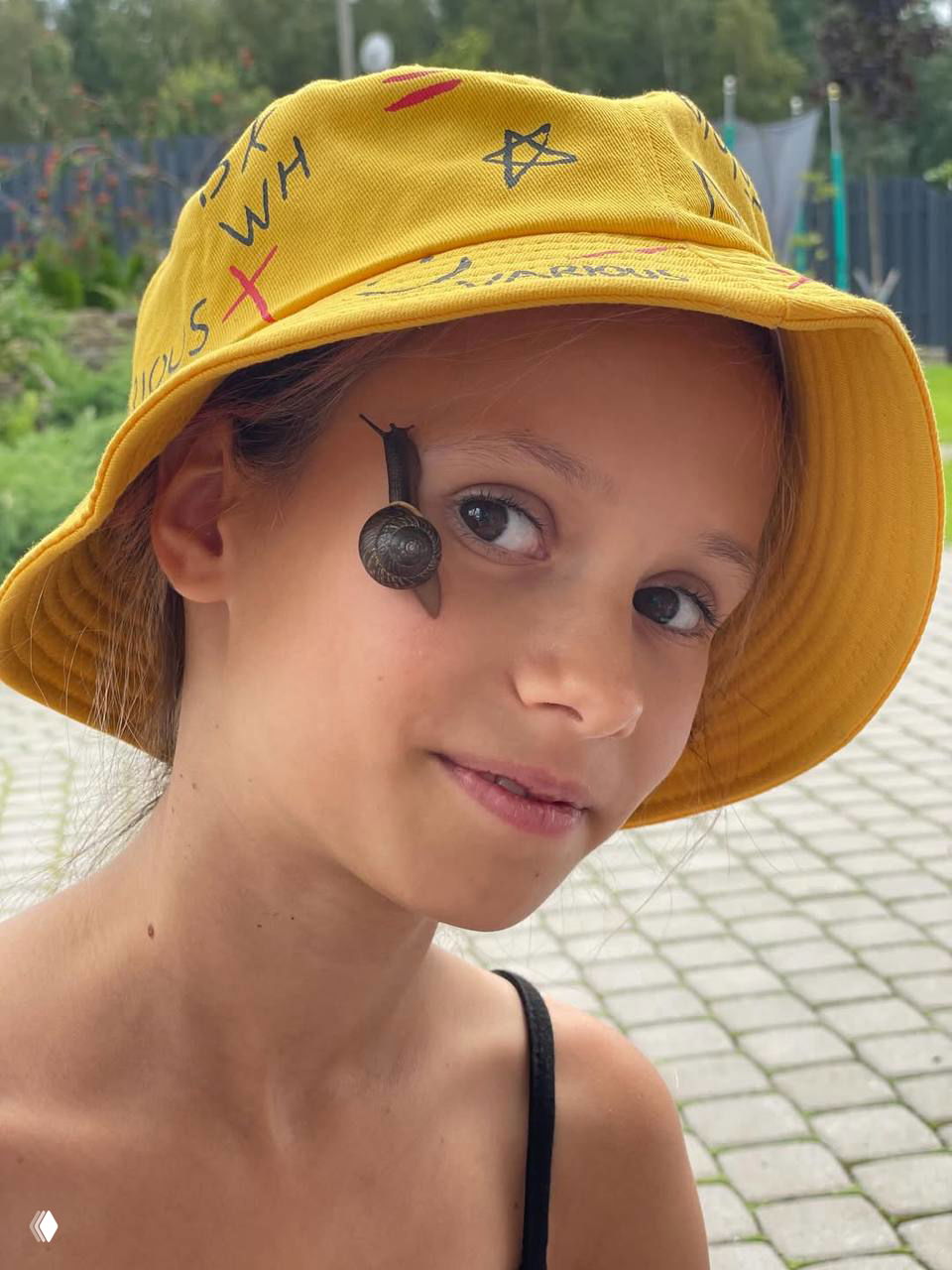 Портрет девочки в желтой панаме на улице; на щеке у нее небольшая улитка — забавный и естественный снимок на прогулке.