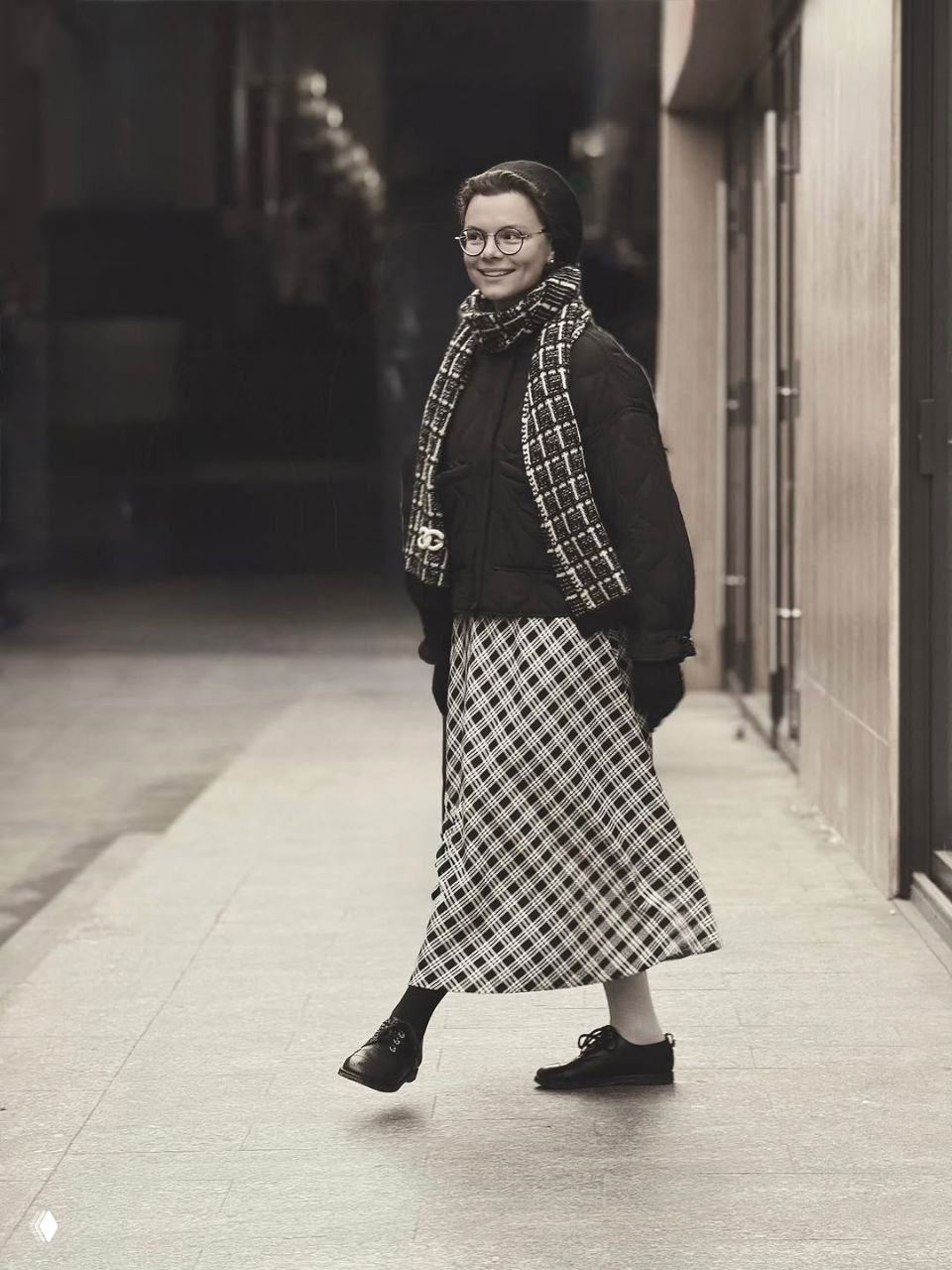 Чёрно‑белый кадр: Татьяна Брухунова в пуховике и клетчатой юбке идёт по улице, эффектный шарф и классические ботинки завершают образ.