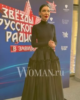 Певица Елка в чёрном платье у баннера фестиваля «Звёзды Русского Радио в Завидово» — снимок с пресс‑зоны с водяным знаком Woman.ru