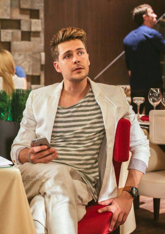 Милош Бикович в светлом костюме в сцене или на съёмочной площадке, сидит за столом с телефоном и внимательным выражением.