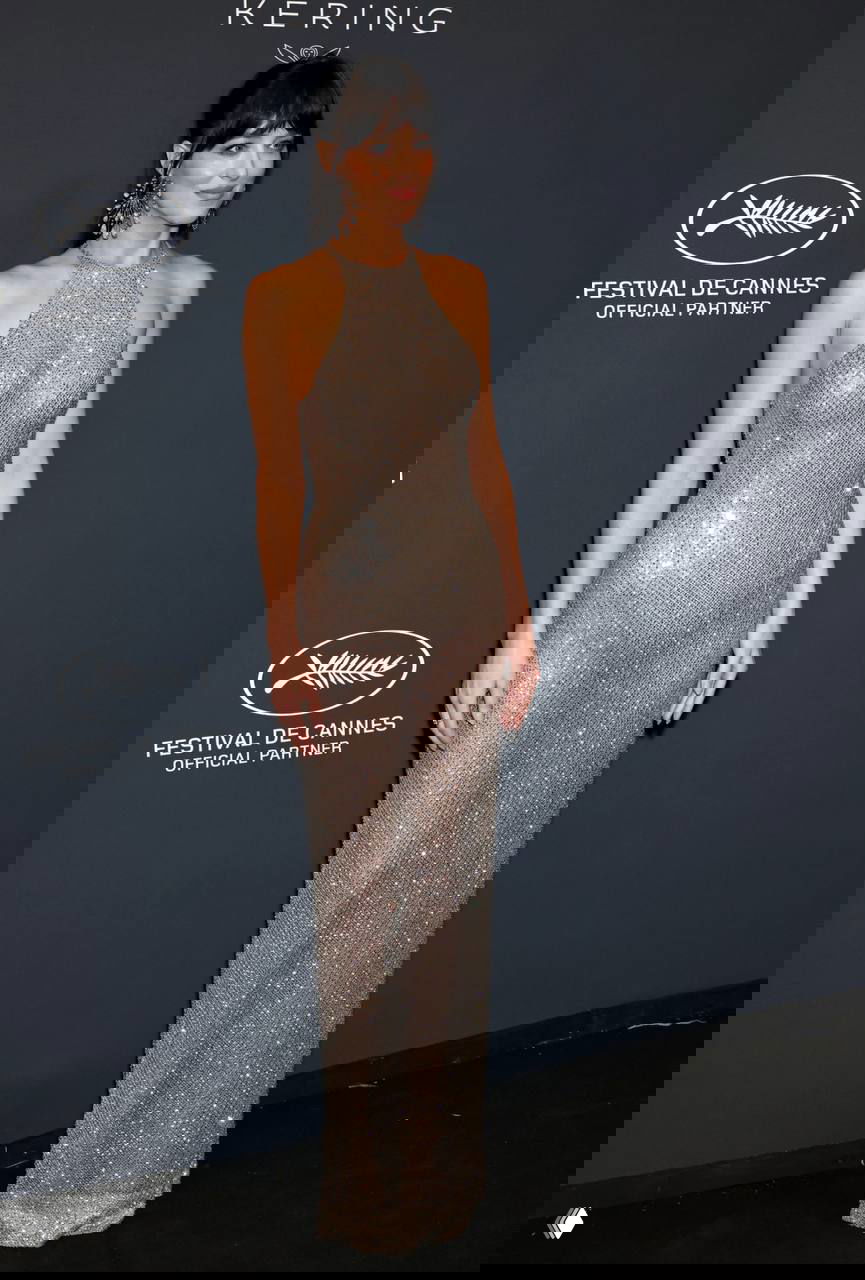 Женщина в облегающем блестящем платье с глубоким вырезом под горлом; украшения и серьги подчёркивают вечерний образ на красной дорожке.