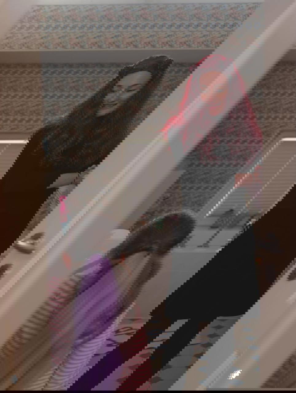 Женщина с тёмно-рыжими волосами улыбается в дверном проёме, рядом ребёнок с фиолетовым хвостиком прически, бытовая обстановка.