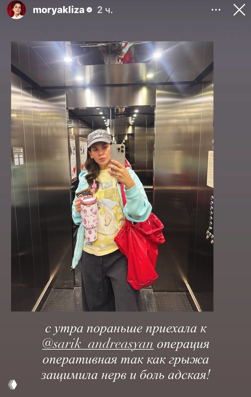 Женщина делает селфи в зеркальном лифте, держит сумку и термокружку; отражения и металлическая отделка лифта хорошо видны на кадре.